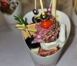 A paper cup contains skewered cured meats, cheese, black grapes, bread, and a sprig of rosemary.