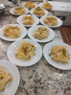 Several plates with layered pastry desserts topped with chopped pistachios and honey are arranged on a granite countertop.