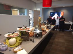 A long table with various buffet dishes and decorations, with four people standing near a TV screen in the background.
