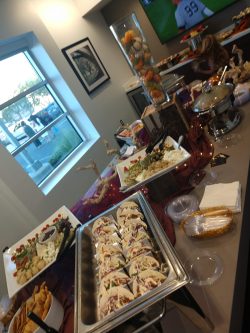 Buffet table with assorted foods, including tacos and salads, set up in a room with a TV showing a football game.