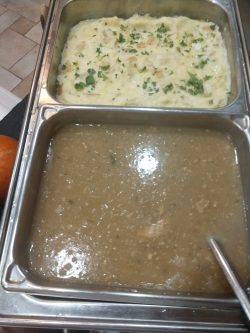 Two metal trays with mashed potatoes topped with herbs above a tray of brown gravy with a serving spoon.
