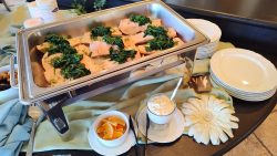 A buffet tray of cooked salmon topped with greens, next to plates, orange slices, and a glass of cream sauce.