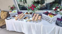 A buffet table with assorted appetizers, fruit, vegetables, shrimp, and flower arrangements displayed on white crates.