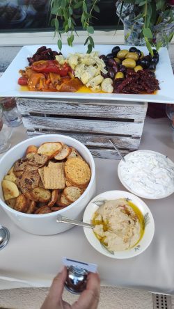A table with a platter of antipasto, a bowl of chips and crackers, hummus, and tzatziki, with a hand holding a condiment.