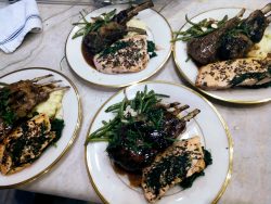 Four plates each with cooked lamb chops, herb-seasoned salmon, mashed potatoes, and green beans on a countertop.