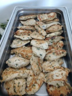 A metal tray filled with grilled, seasoned chicken breast fillets arranged in overlapping layers.