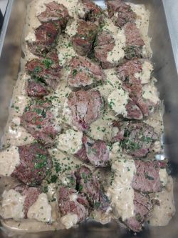 Sliced beef medallions topped with creamy sauce and chopped herbs in a stainless steel tray.