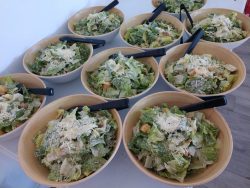 Ten wooden bowls of Caesar salad with lettuce, croutons, cheese, and black-handled tongs arranged on a table.