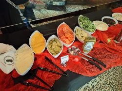 A buffet table with sliced cheese, pickles, tomatoes, lettuce, sauces, and utensils arranged on red cloth.