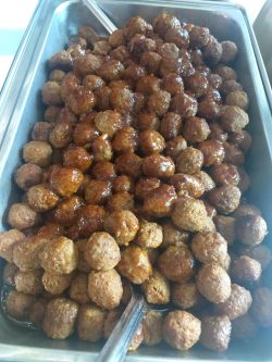 A large metal tray filled with glazed meatballs, with serving tongs placed at the bottom left corner.