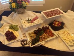 A table with assorted cheeses, grapes, vegetables, crackers, meats, and a floral arrangement in the background.