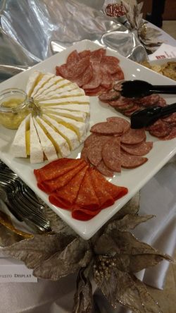 A platter with sliced brie cheese, assorted cured meats, a small jar of mustard, and serving tongs on a table.