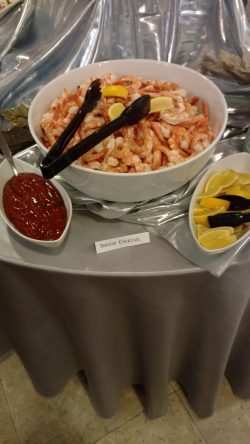 A bowl of shrimp cocktail with lemon wedges and cocktail sauce on the side, sitting on a table with tongs.