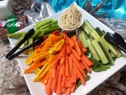 A platter of sliced carrots, cucumbers, celery, bell peppers, and dip, arranged with tongs on a white square plate.