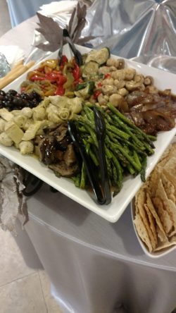 A platter of assorted roasted vegetables and olives with serving tongs next to a stack of flatbread on a table.