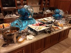 Buffet table with various appetizers, meatballs, cold cuts, and blue balloon centerpieces in a kitchen setting.